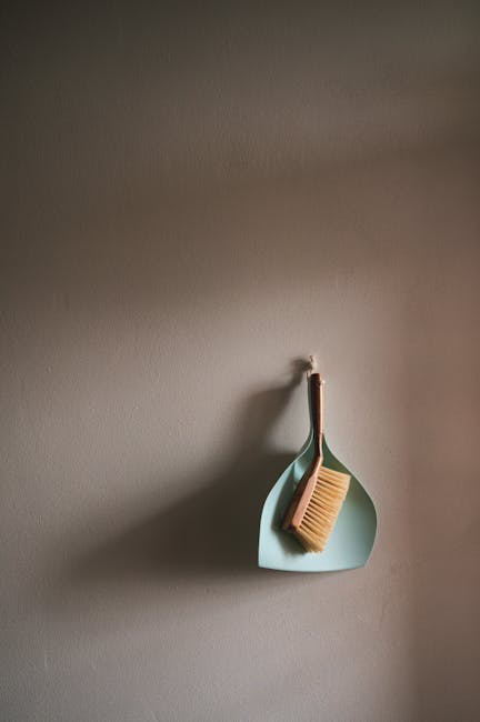 A light mint green dustpan and a wooden-handled cleaning brush with beige bristles hanging on a white hook against a smooth, neutral grey wall. The dustpan and brush are clean and well-maintained, wit