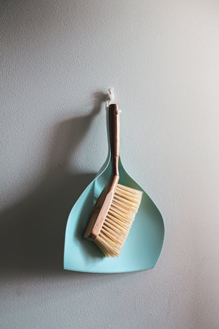 A cleaning brush with light wood handle and beige bristles hangs on a white wall, inside a light blue dustpan with a curved shape. The dustpan and brush are positioned near each other, with the handle
