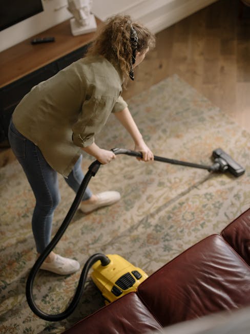 A close-up view of a modern vacuum cleaner with a transparent dust container filled with dust and debris, connected to a flexible, patterned hose that extends to a black floor attachment in a domestic